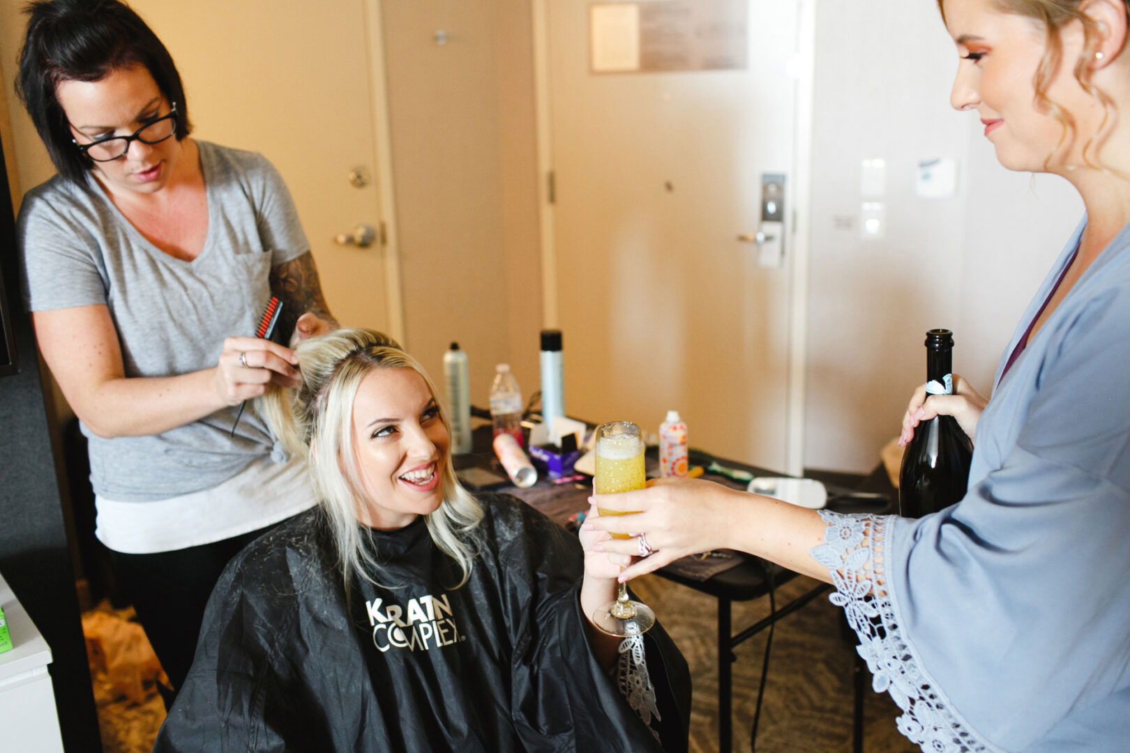 At the French Creek Golf Club Wedding, a woman enjoys a drink while having her hair styled. Nearby, an array of hair products and styling tools await their turn to perfect her look for the special day.