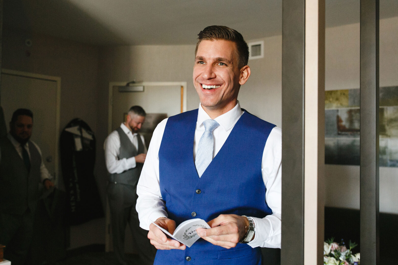 A man in a blue vest and white shirt smiles while holding a card at a French Creek Golf Club wedding, with two other men in the background.