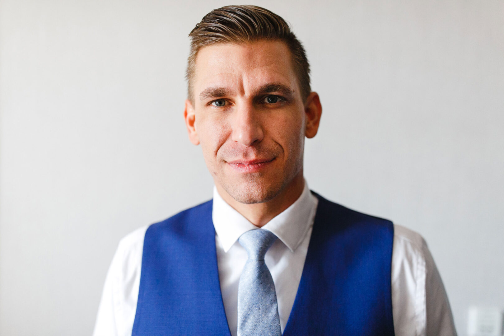 Man in a white shirt and blue vest, wearing a light blue tie, gazes at the camera against a plain background, reminiscent of elegance seen at French Creek Golf Club weddings.