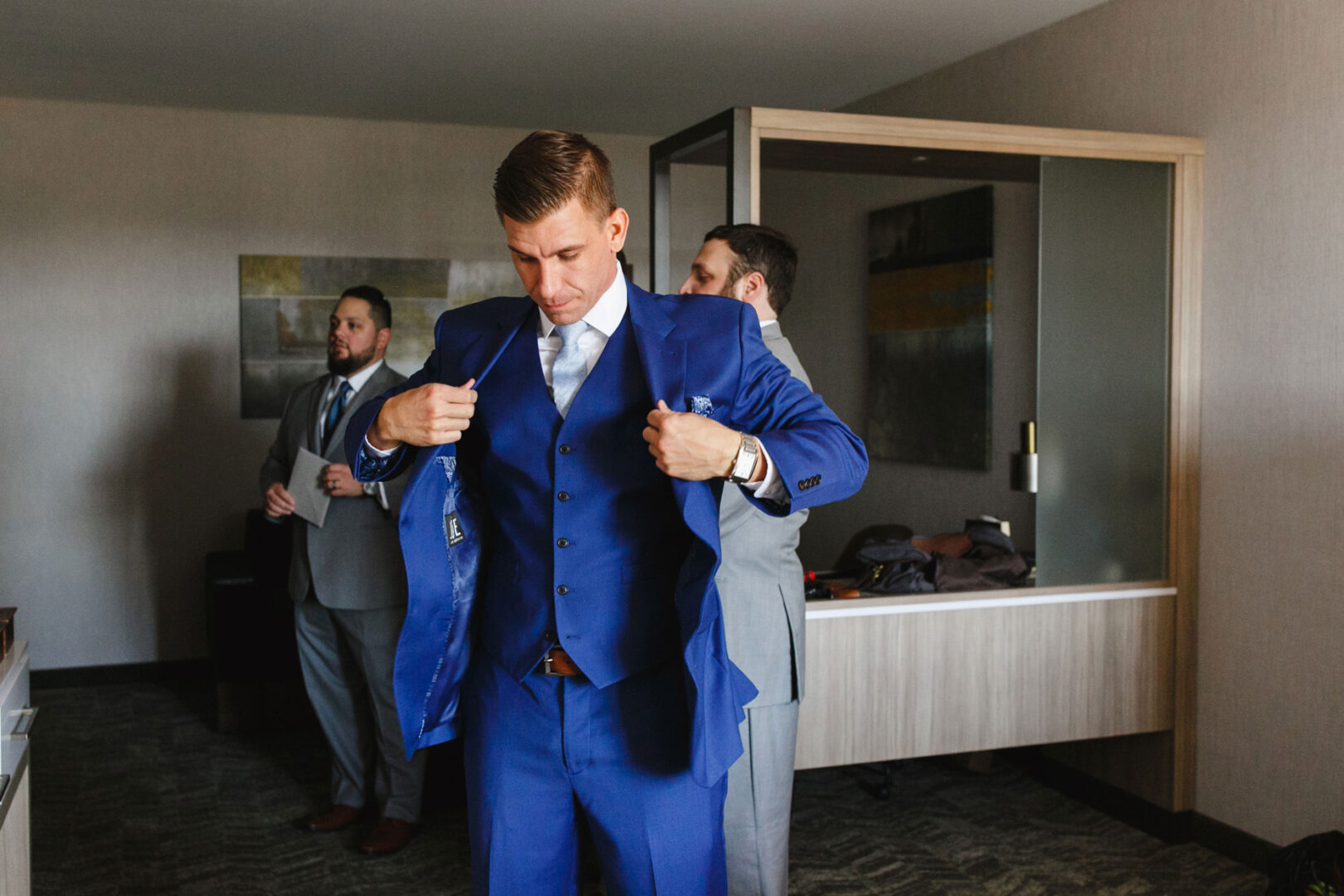A man in a blue suit adjusts his jacket while another stands behind him, preparing for the French Creek Golf Club wedding. A third man in a gray suit stands in the background, all set within a room boasting modern decor.