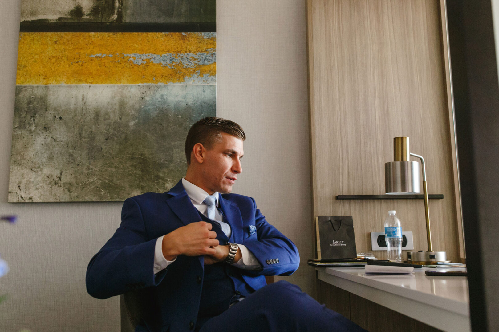 A man in a blue suit sits at a desk adjusting his jacket, as if preparing for an event as elegant as a French Creek Golf Club wedding. In the background, a lamp, water bottle, and abstract painting add to the sophisticated ambiance.
