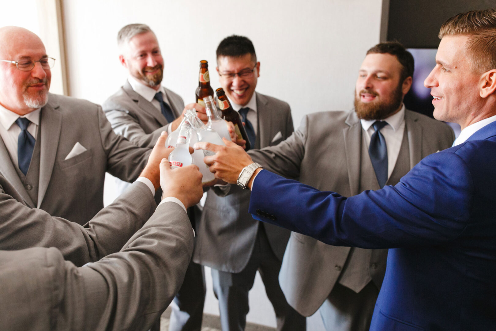 A group of six men dressed in suits clink bottles in a celebratory toast at the picturesque French Creek Golf Club wedding.