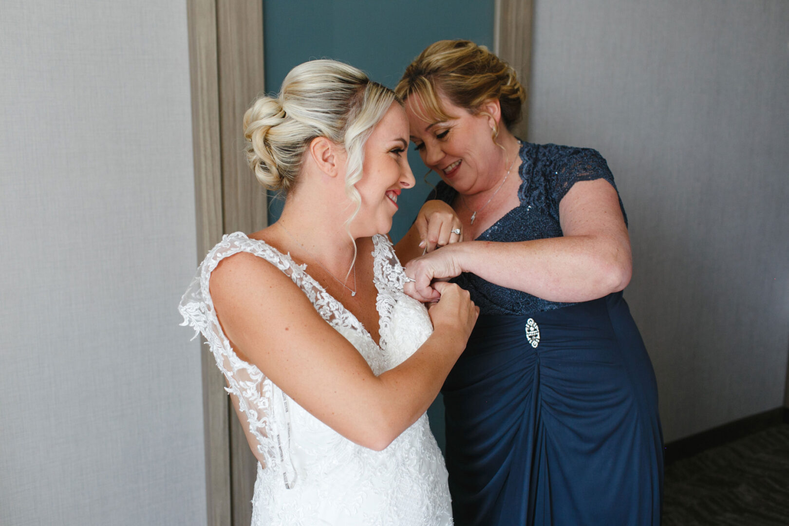Inside a room at the French Creek Golf Club, a bride in a lace wedding dress is being gently assisted by a woman in a blue dress, preparing for her special day.