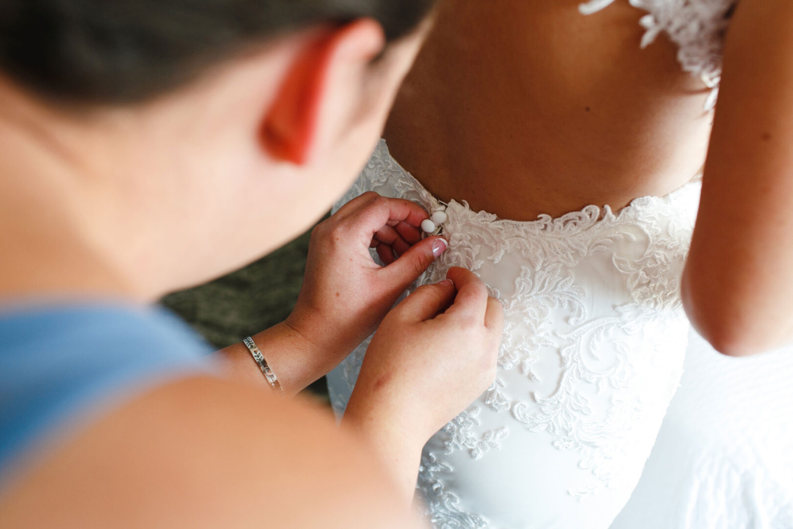 At a French Creek Golf Club wedding, delicate hands carefully button the back of an elegant white lacy wedding dress worn by a radiant bride.