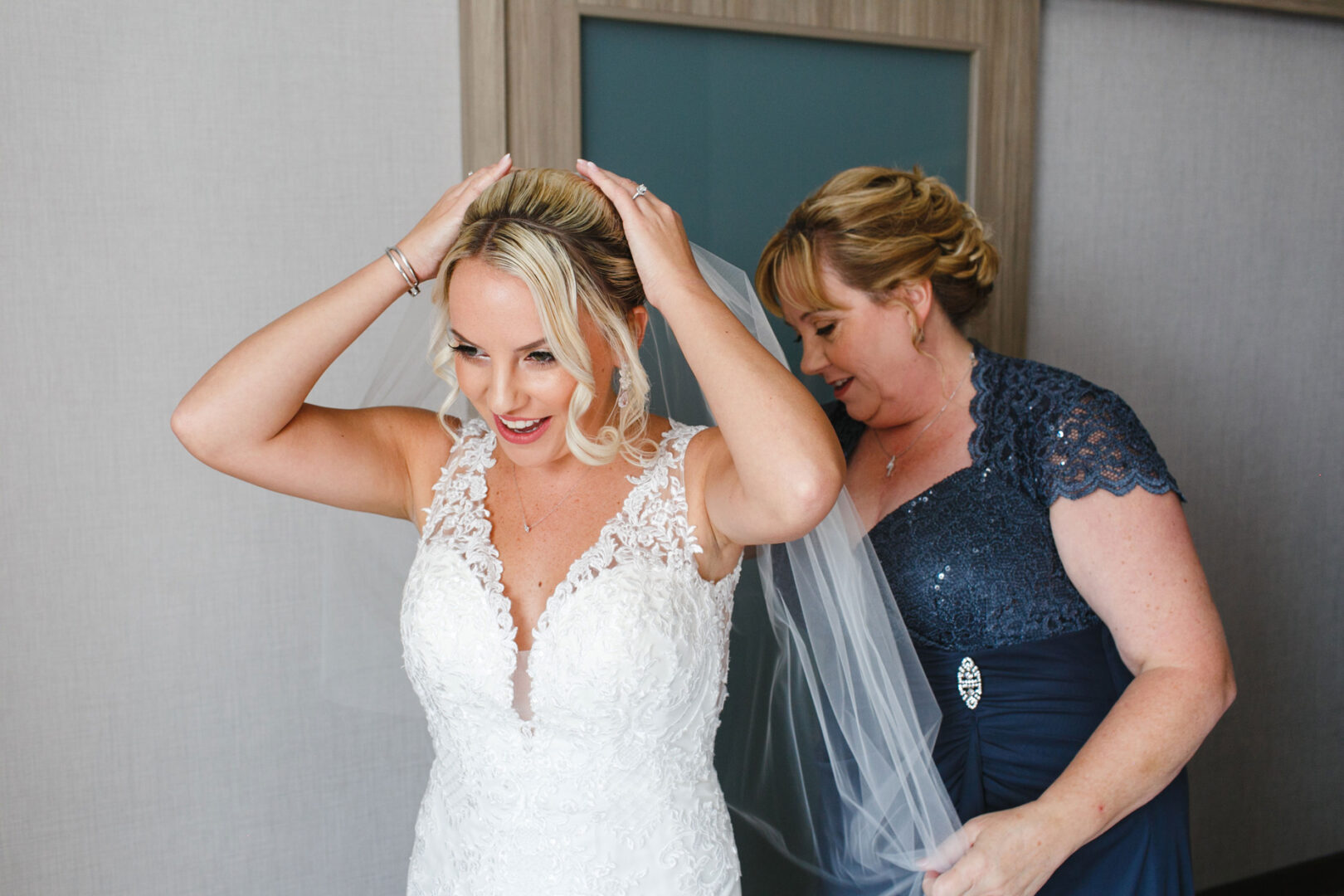 In a room at the French Creek Golf Club, a bride in a white lace dress adjusts her hair while a woman in a blue dress assists with the veil.