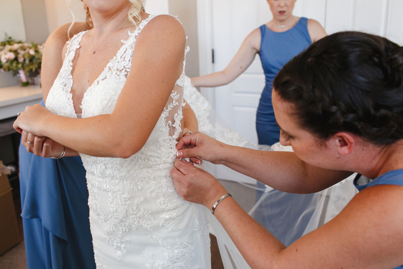A bride in a stunning white lace wedding dress is being assisted with the back of her gown by a woman at a charming French Creek Golf Club wedding. Other women in elegant blue dresses are also present, adding to the joyous occasion.