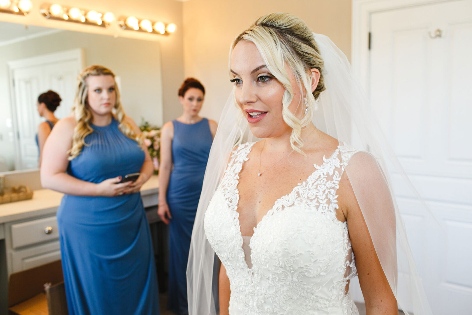 In a brightly lit room at the French Creek Golf Club Wedding, a bride in a white dress and veil stands gracefully, with her two bridesmaids in blue dresses elegantly poised in the background.