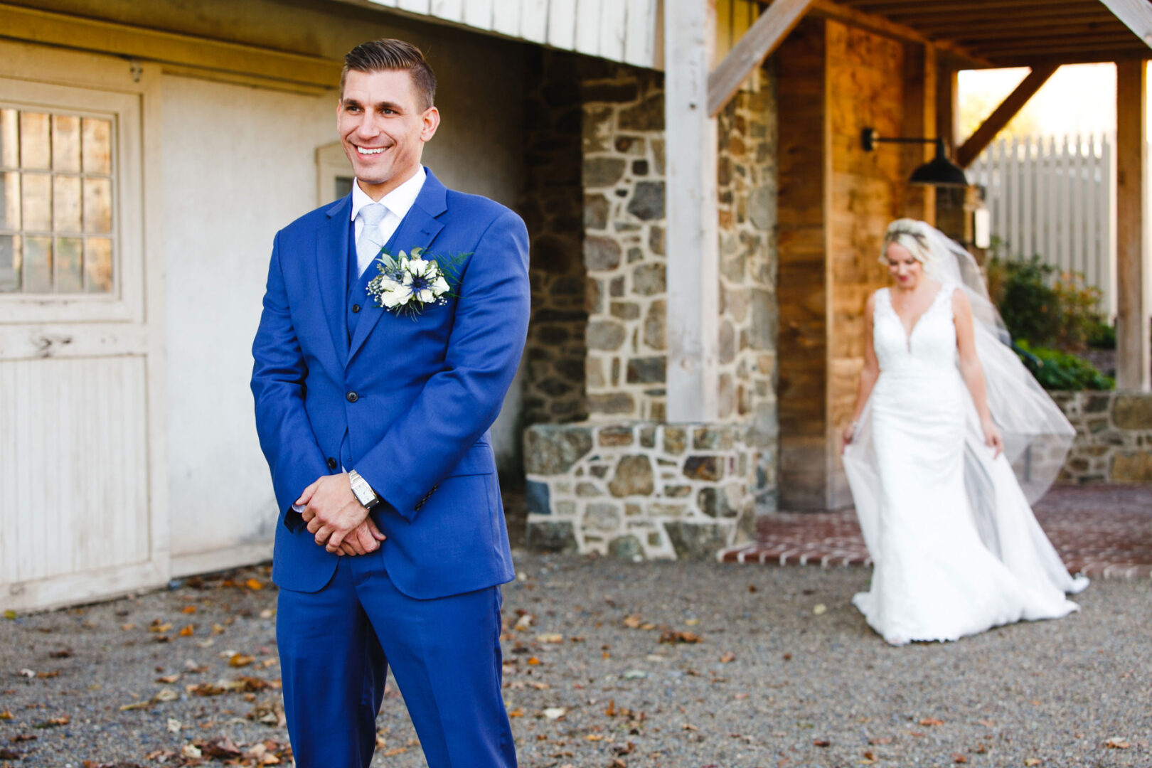 Groom in a blue suit stands smiling outdoors at the French Creek Golf Club Wedding, as a bride in a white gown approaches from behind near a stone wall and wooden structure.