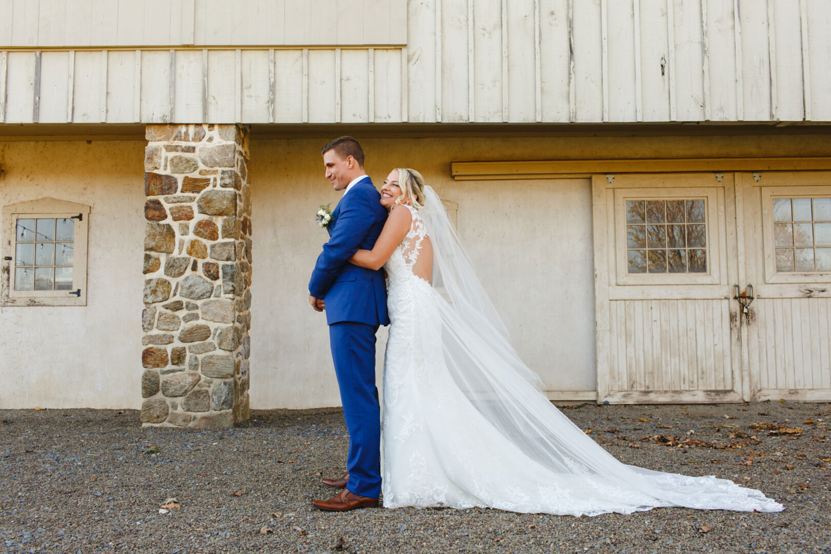 At a charming French Creek Golf Club wedding, a bride in a white gown tenderly embraces her groom, who is dressed in a blue suit. They stand lovingly before a rustic stone and wood building.