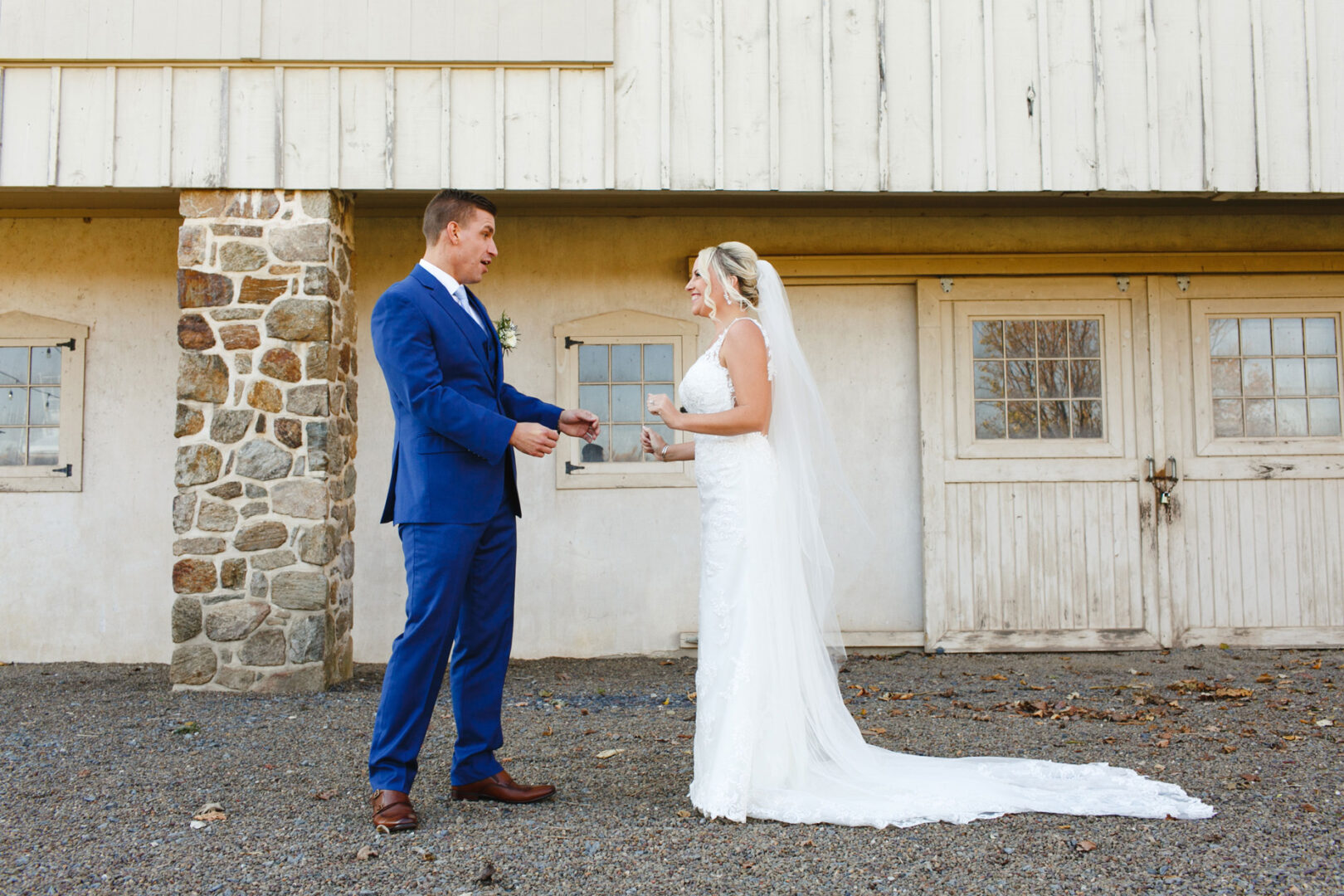 The bride and groom stand facing each other outside a rustic building at the picturesque French Creek Golf Club. The groom is in a blue suit, while the bride dazzles in a white gown with a long veil, creating an enchanting wedding scene.
