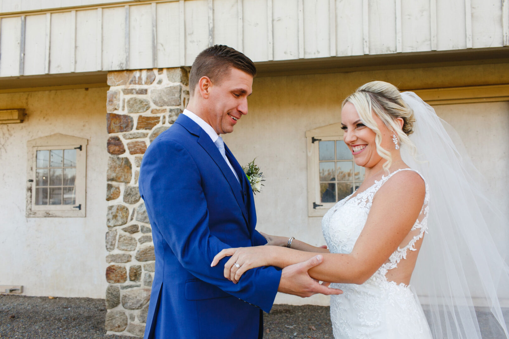 The bride and groom, holding hands and smiling warmly at each other outside a building with stone pillars, celebrate their French Creek Golf Club wedding. The groom looks dapper in his blue suit, while the bride dazzles in her white gown with a veil.