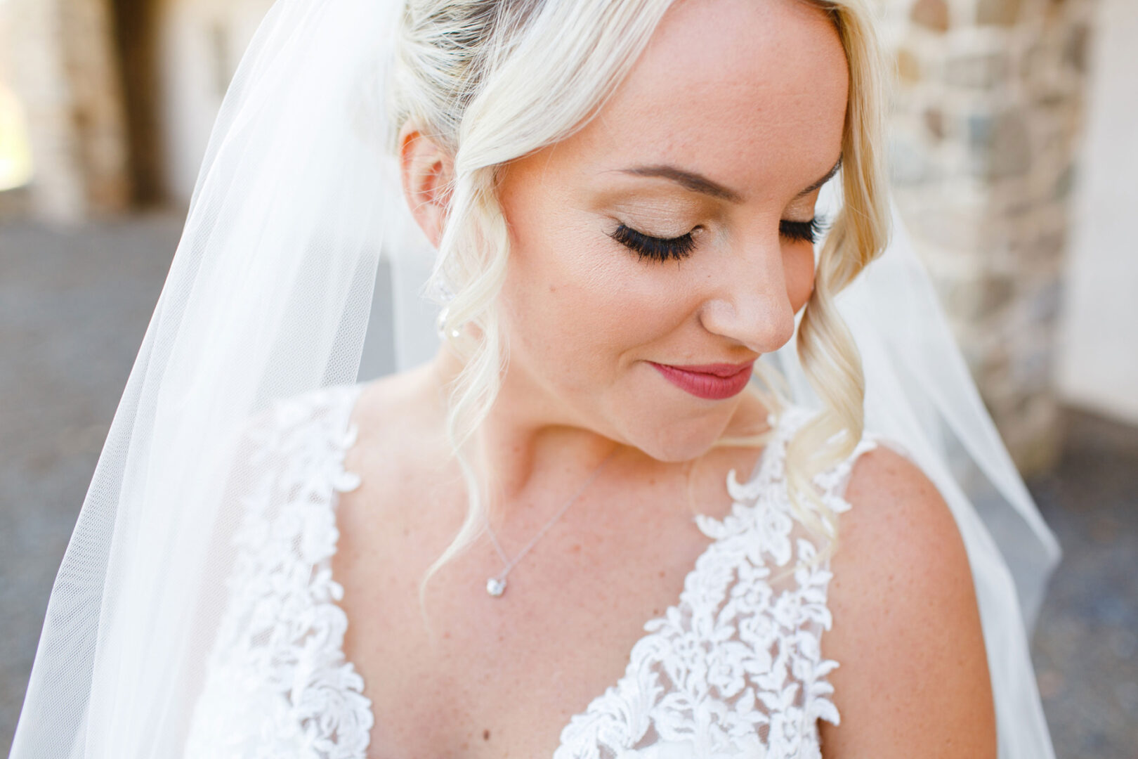 The bride, with her blonde hair and lace dress, smiles softly as she gazes down. She wears a delicate veil and subtle makeup, embodying elegance against the backdrop of stone and blurred architectural elements at the enchanting French Creek Golf Club Wedding.