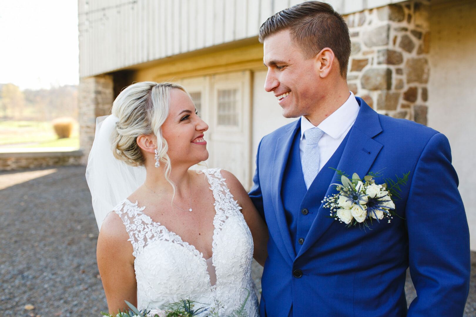 The bride and groom stand smiling at each other, with the bride in a white dress and veil and the groom in a blue suit adorned with a boutonniere. They are outside near a stone building, capturing the elegance of their French Creek Golf Club wedding.