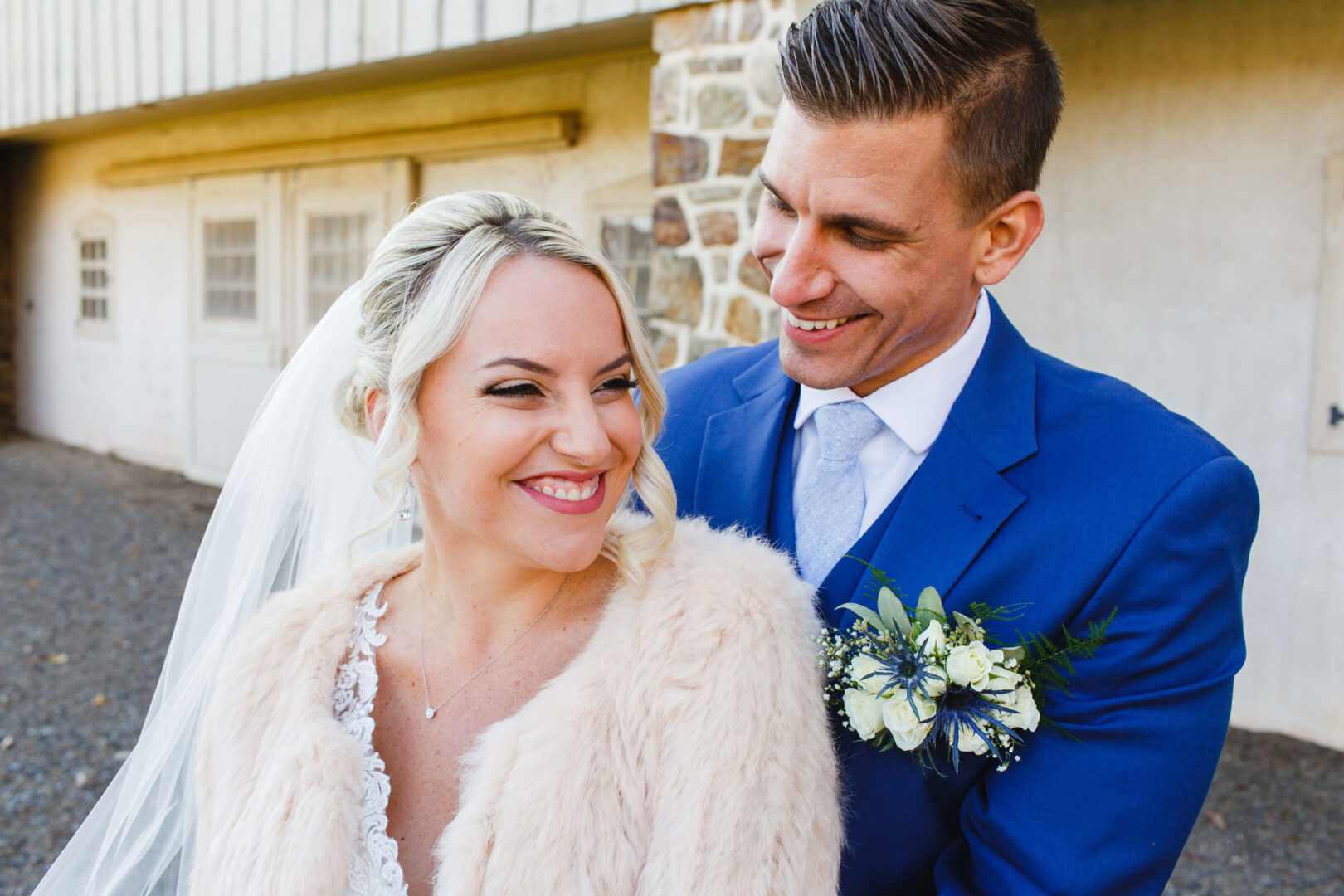 A bride and groom stand together smiling at their French Creek Golf Club wedding. The bride wears a fur shawl and veil, while the groom is in a blue suit with a floral boutonniere. They grace an outdoor setting framed by rustic stone walls.
