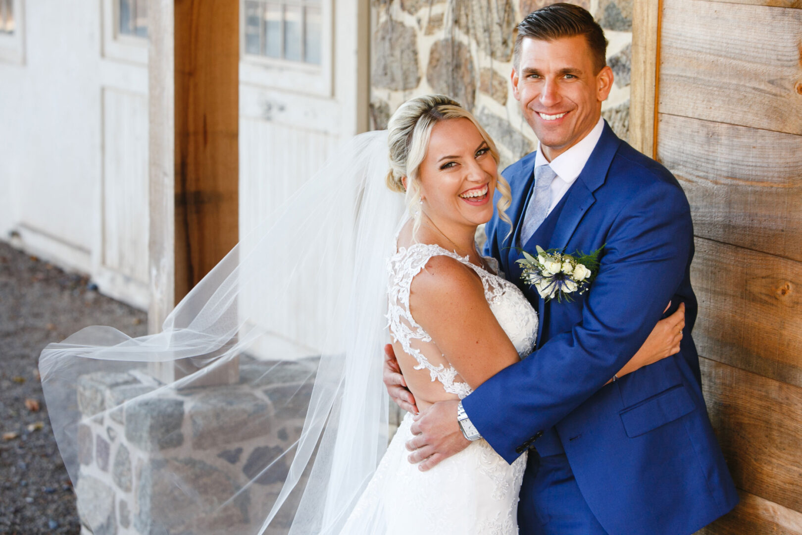Bride and groom smiling and posing together outdoors near a rustic building at their French Creek Golf Club wedding. The bride is in a white gown and veil, while the groom is in a blue suit.