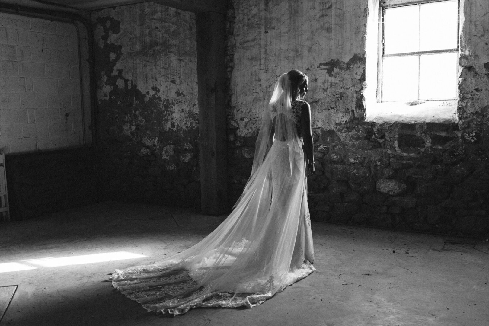 The bride in her long gown and veil stands alone in a dimly lit, rustic room at the French Creek Golf Club Wedding, with a stone wall and window, gazing out.