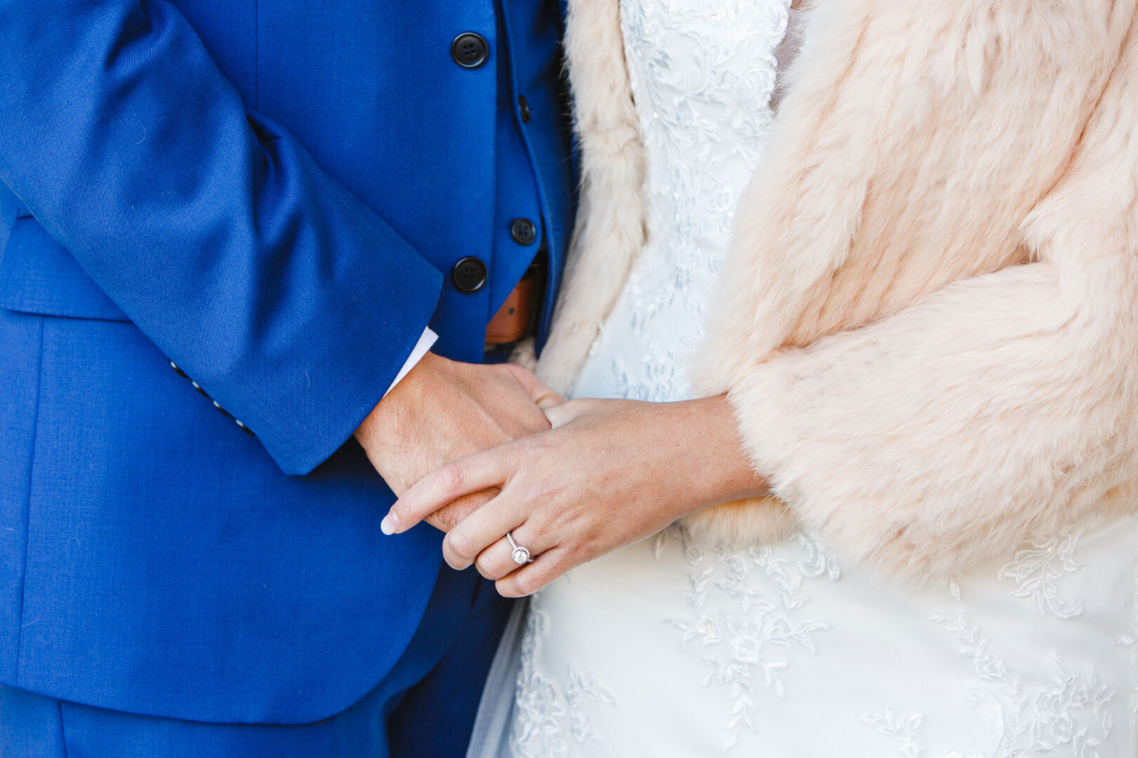 At the French Creek Golf Club wedding, a couple holds hands; one wears a blue suit, and the other is in a white lace dress with a beige fur stole.