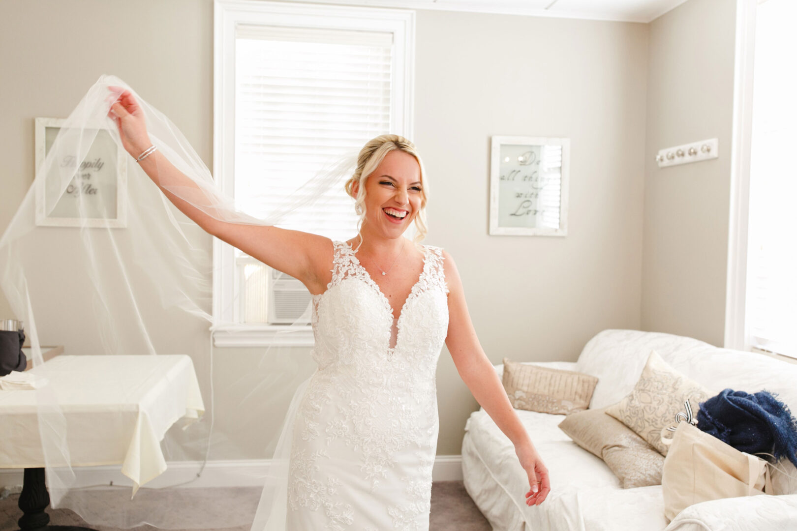 Bride in a white lace wedding dress holds a veil, smiling in a bright, cozy room with soft furnishings and framed wall art at her French Creek Golf Club Wedding.
