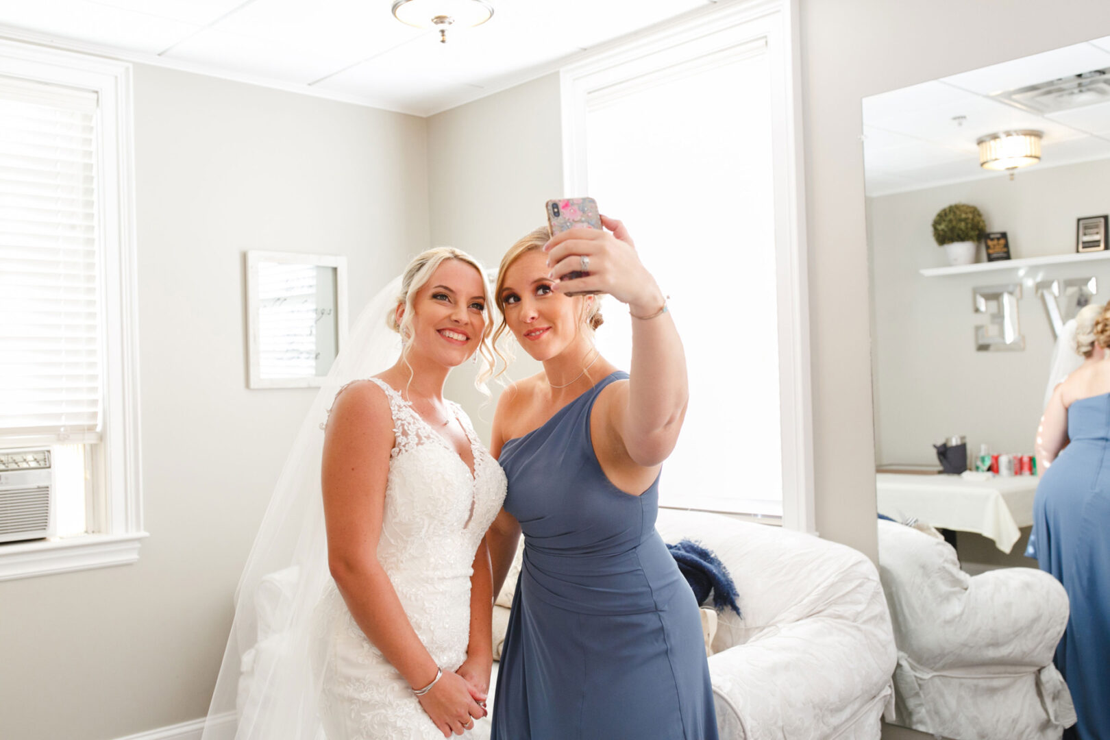 Two women in a room capturing a selfie; one dons a white wedding dress, embodying the elegance of a French Creek Golf Club wedding, while the other shines in a blue dress.