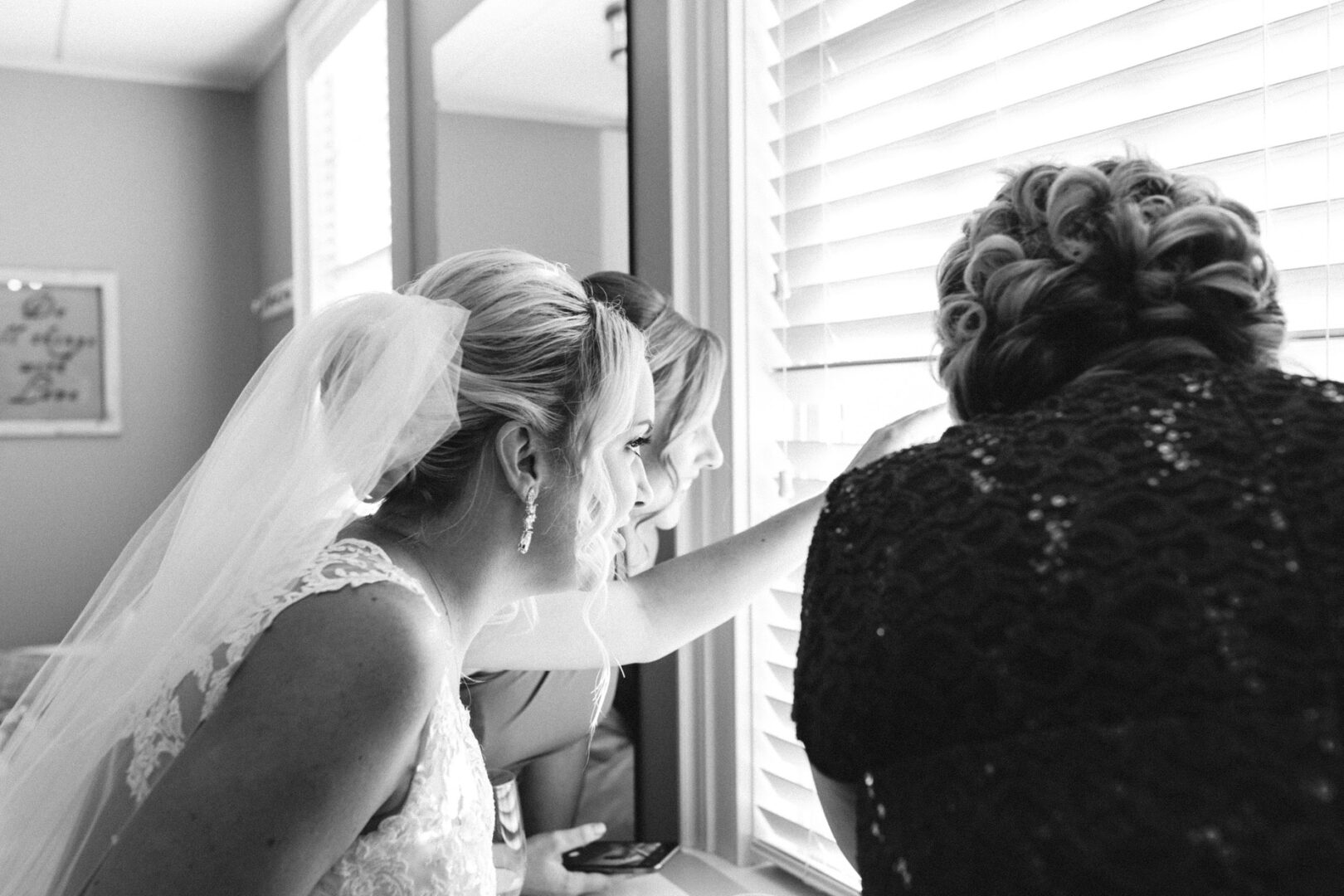 Three women in formal attire gaze through the window blinds at the French Creek Golf Club wedding. The bride, draped in a veil on the left, clutches her phone. This elegant black and white image captures a timeless moment of anticipation.