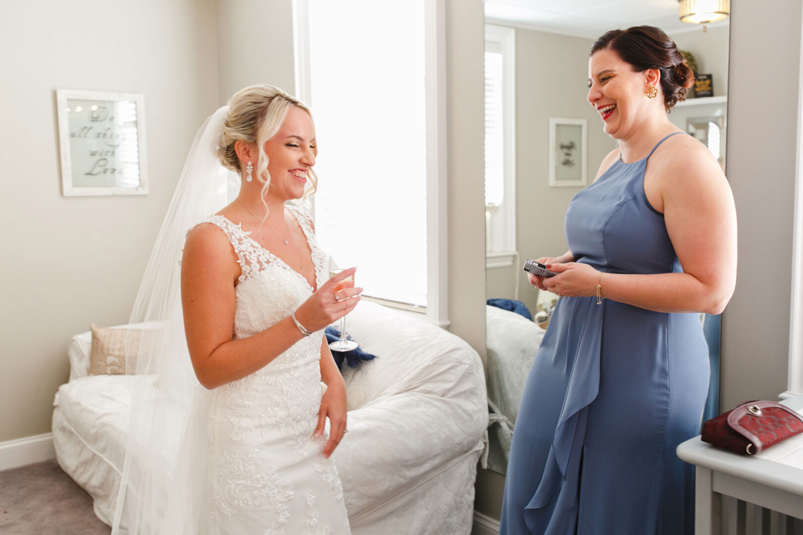 In the sunlit room of the French Creek Golf Club, a bride in a white dress adorned with a veil shares a laugh with her bridesmaid in a stunning blue gown, capturing the essence of joy and elegance on this special day.