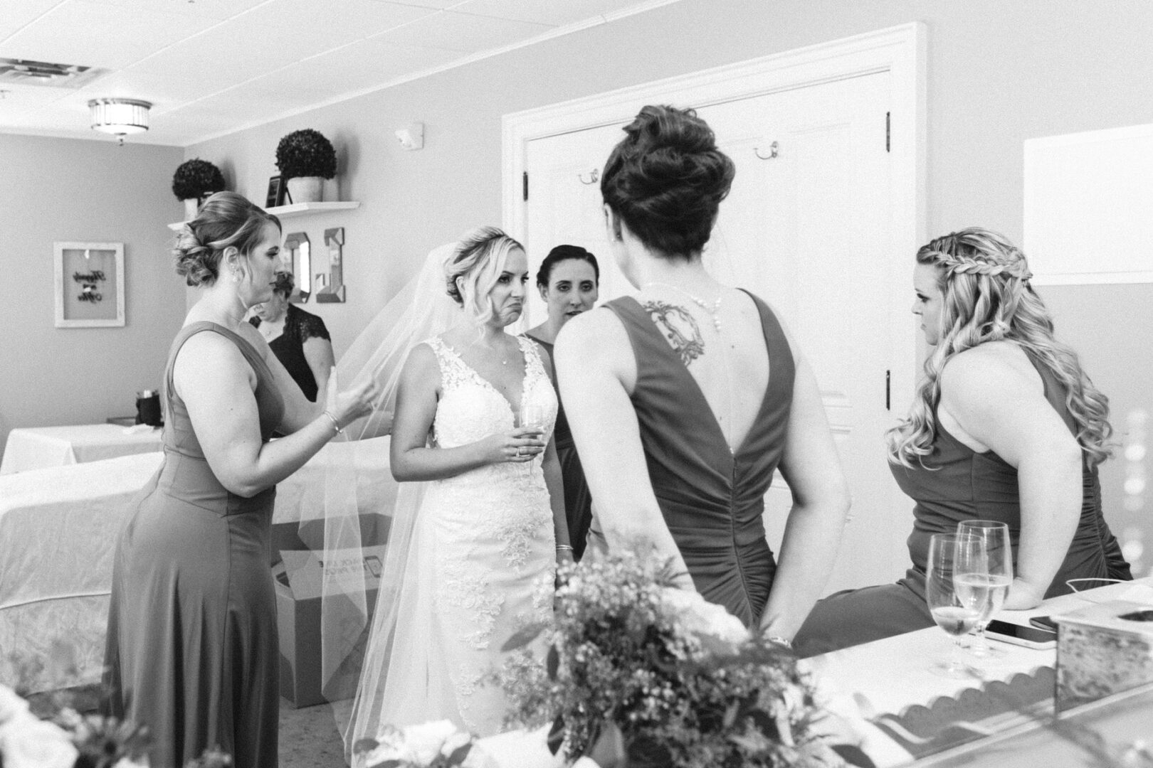 The bride, in her elegant wedding dress and veil, stands with her bridesmaids at the French Creek Golf Club Wedding. They are dressed in formal gowns, engaging in lively conversation. Nearby, tables are set neatly with glasses of water sparkling under the light.