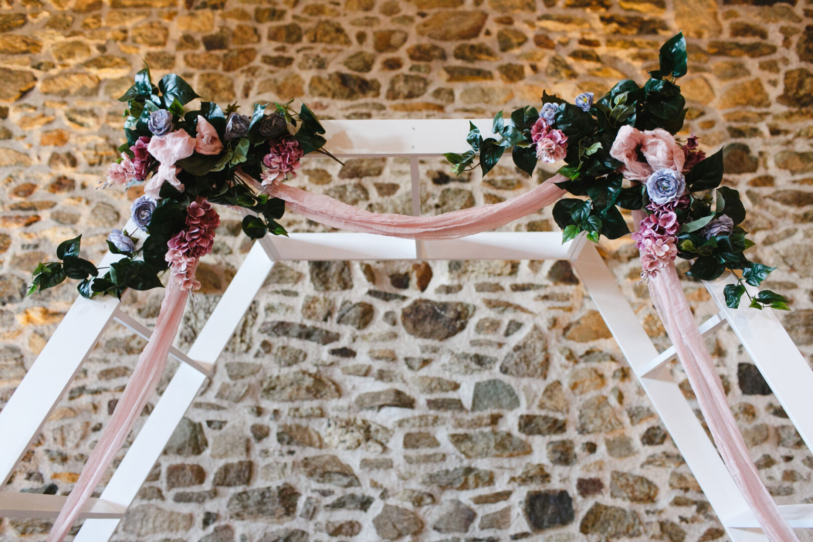 A stunning wooden arch adorned with pink and purple flowers and lush green leaves stands elegantly against the stone wall backdrop, setting the perfect scene for a French Creek Golf Club wedding.