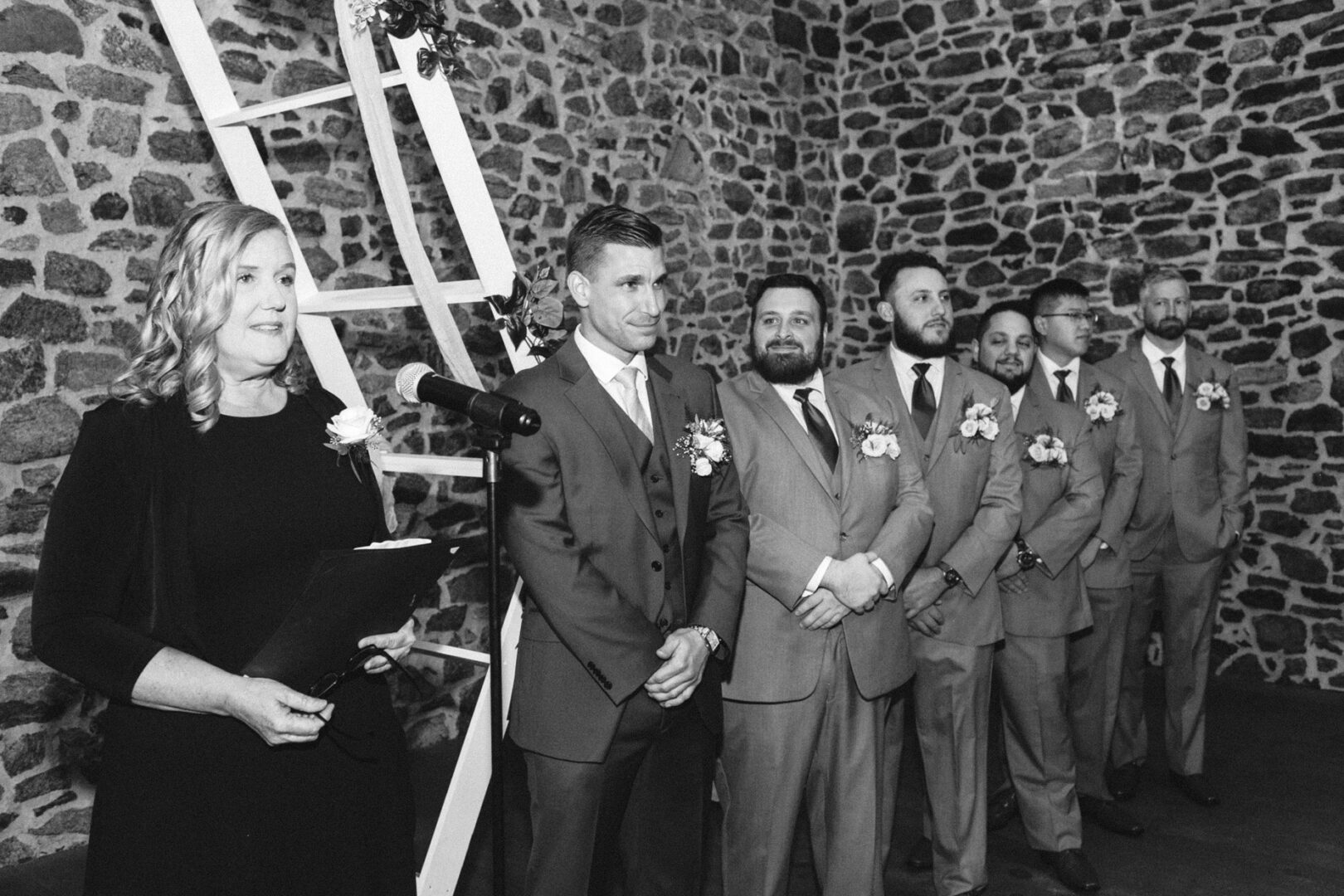 A woman stands at a microphone holding papers, facing a line of men in suits and ties with boutonnieres, against the rustic charm of the stone wall at the French Creek Golf Club wedding.