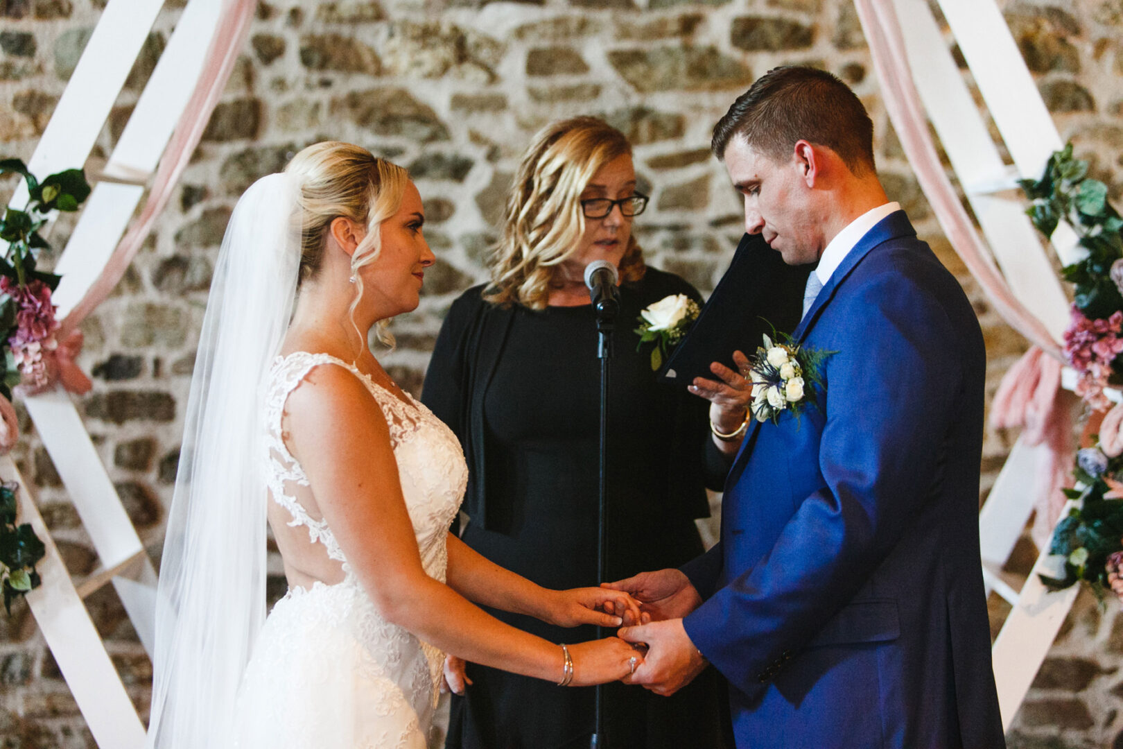 During their French Creek Golf Club wedding, a couple exchanges rings before an officiant. The bride dazzles in a white dress with a veil, while the groom looks sharp in a blue suit. They stand poised before a stone wall and floral arch.