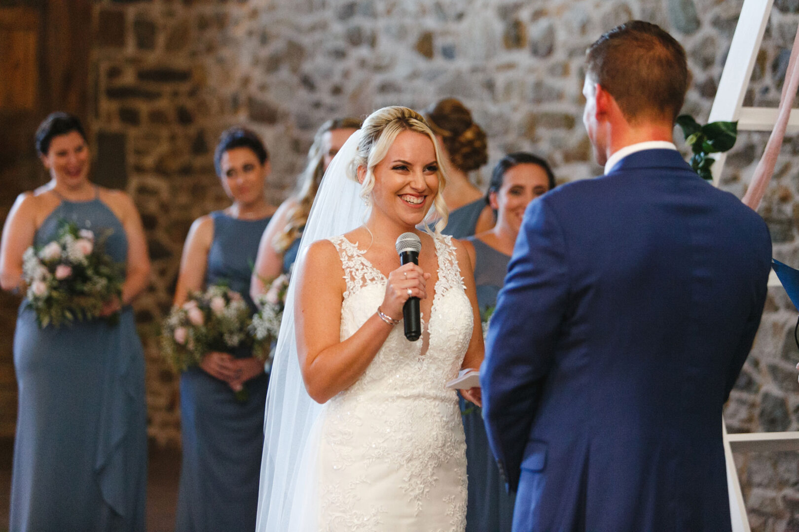 At a beautiful French Creek Golf Club wedding, a bride in a white dress and veil speaks into a microphone, facing a groom in a blue suit. Bridesmaids in blue dresses hold bouquets elegantly in the background, making the moment unforgettable.