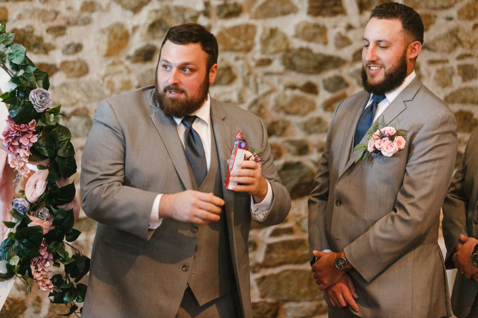 Two men in gray suits stand by a stone wall at the French Creek Golf Club wedding. One holds a can wrapped in his hand, while the other looks on, smiling. They are surrounded by floral decorations.