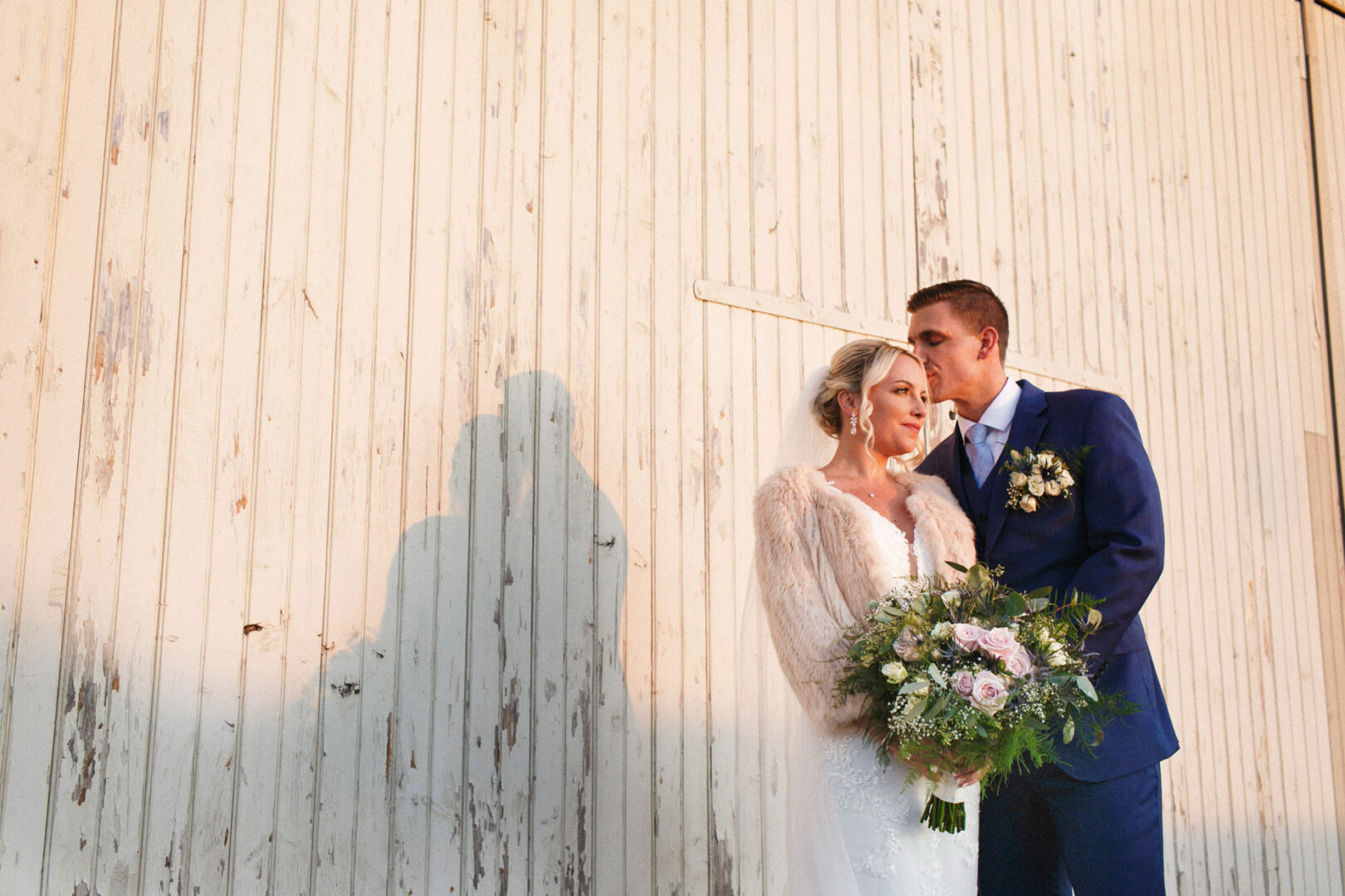 The bride and groom stand close together, holding a bouquet, in front of a rustic wooden wall at their enchanting French Creek Golf Club wedding.