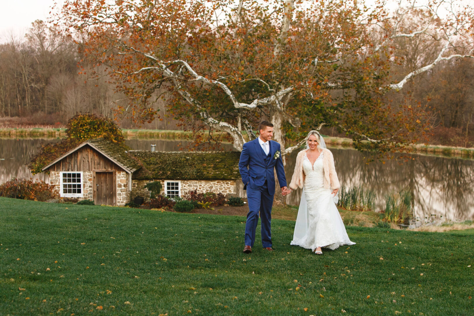 A bride and groom walk hand in hand on a grassy area at French Creek Golf Club, near a small stone building and a serene lake, with autumn trees in the background.