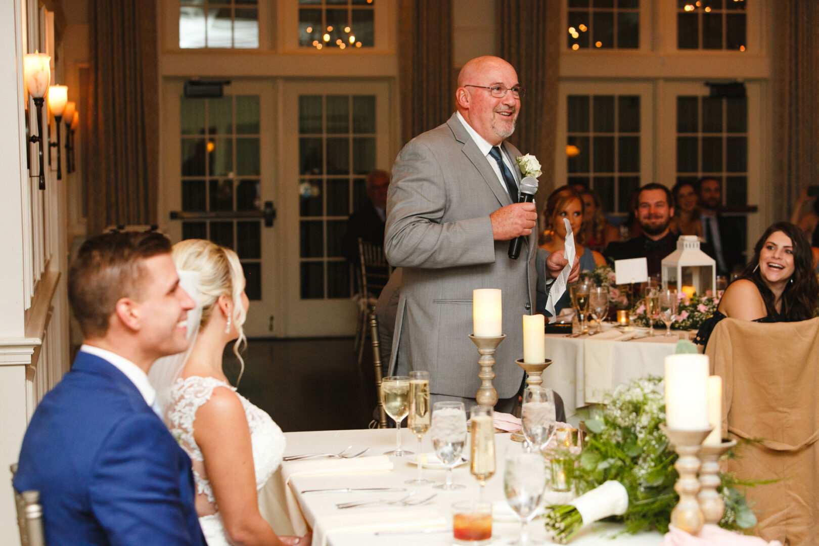 A man in a gray suit speaks into a microphone at the French Creek Golf Club wedding reception. The bride and groom sit at a table decorated with candles and greenery, while guests listen in the background.