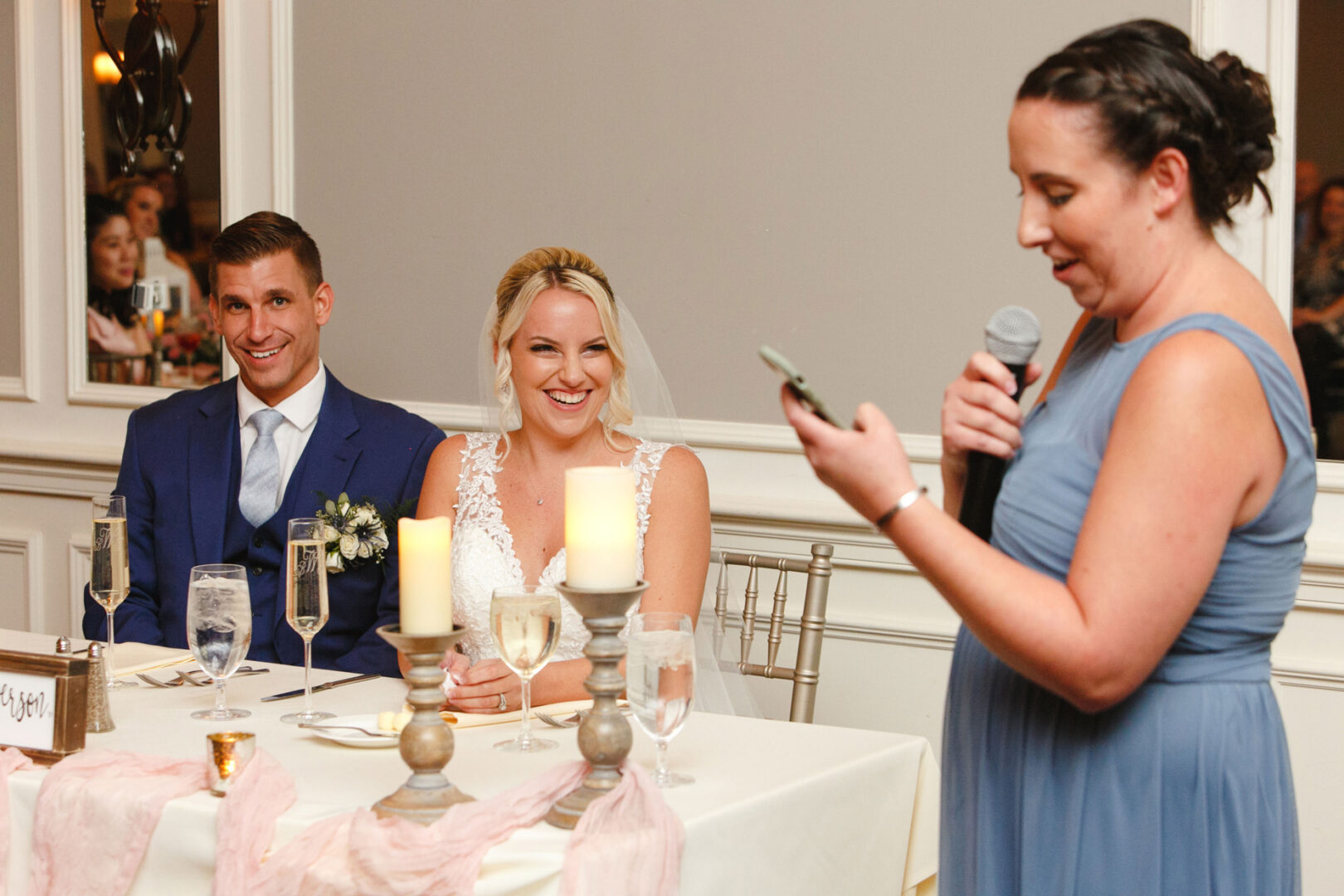 At a beautiful French Creek Golf Club wedding, a person in a blue dress delivers a heartfelt speech. The couple, elegantly attired in a suit and wedding gown, sit amidst candles and decorations, listening and smiling warmly.