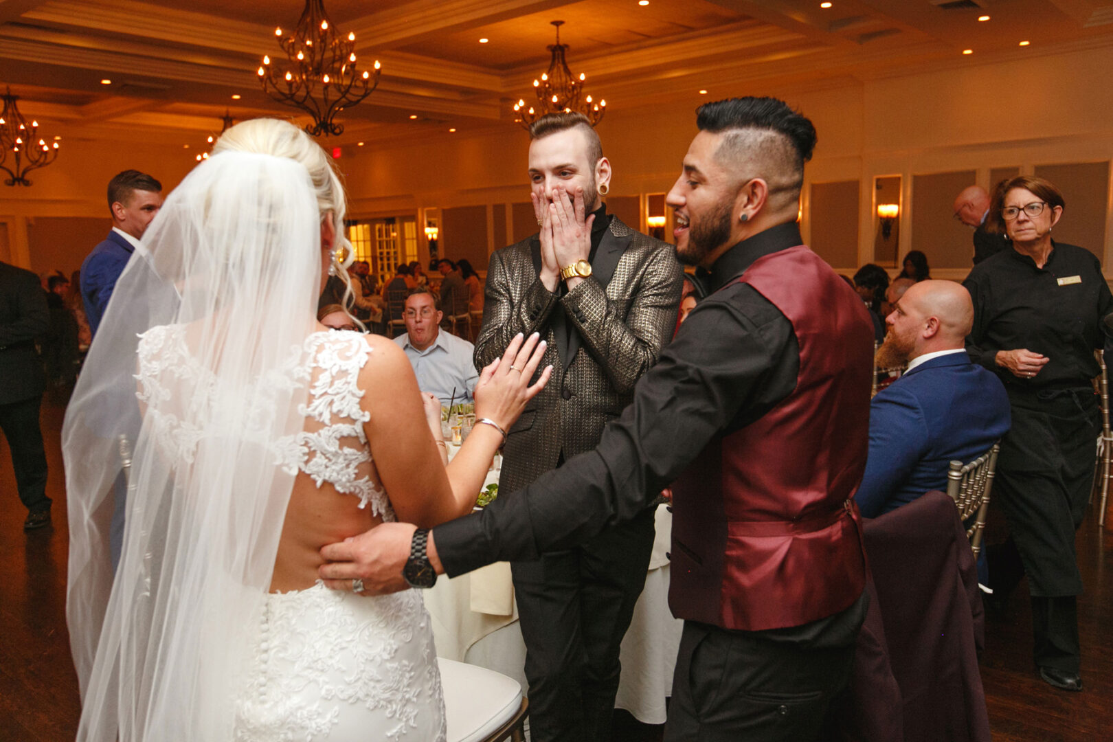 At a French Creek Golf Club wedding, a bride in a lace gown and veil engages with two men at the formal event. One man gestures with surprise near his face, while the other, in a dark vest, smiles warmly. A chandelier's glow illuminates the scene.
