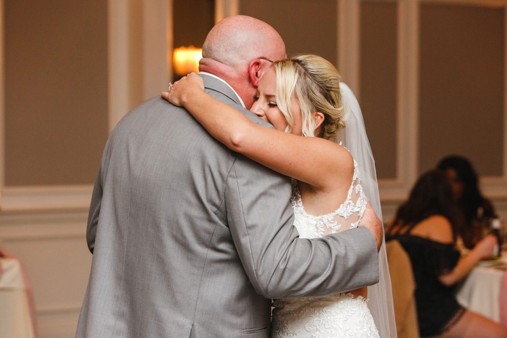 In a warmly lit room at French Creek Golf Club, a bride in a white lace dress embraces an older man in a gray suit, both appearing emotional.