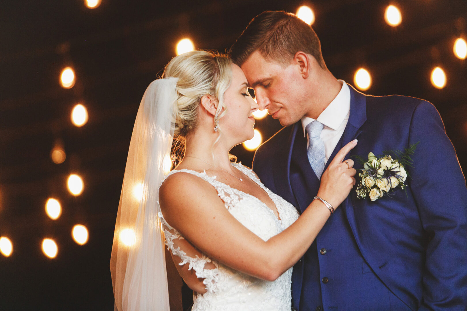 Bride and groom embrace under warm string lights at their French Creek Golf Club wedding. She wears a lace dress; he wears a blue suit with a floral boutonniere.