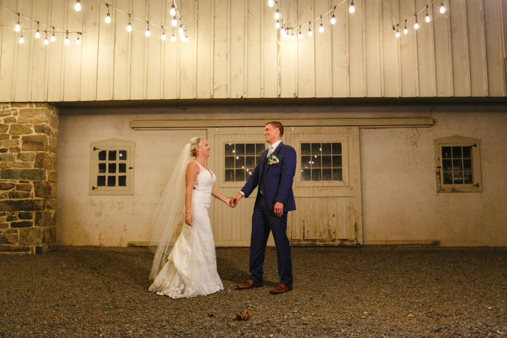 Bride and groom hold hands, standing under string lights in front of a barn with stone walls at the French Creek Golf Club.