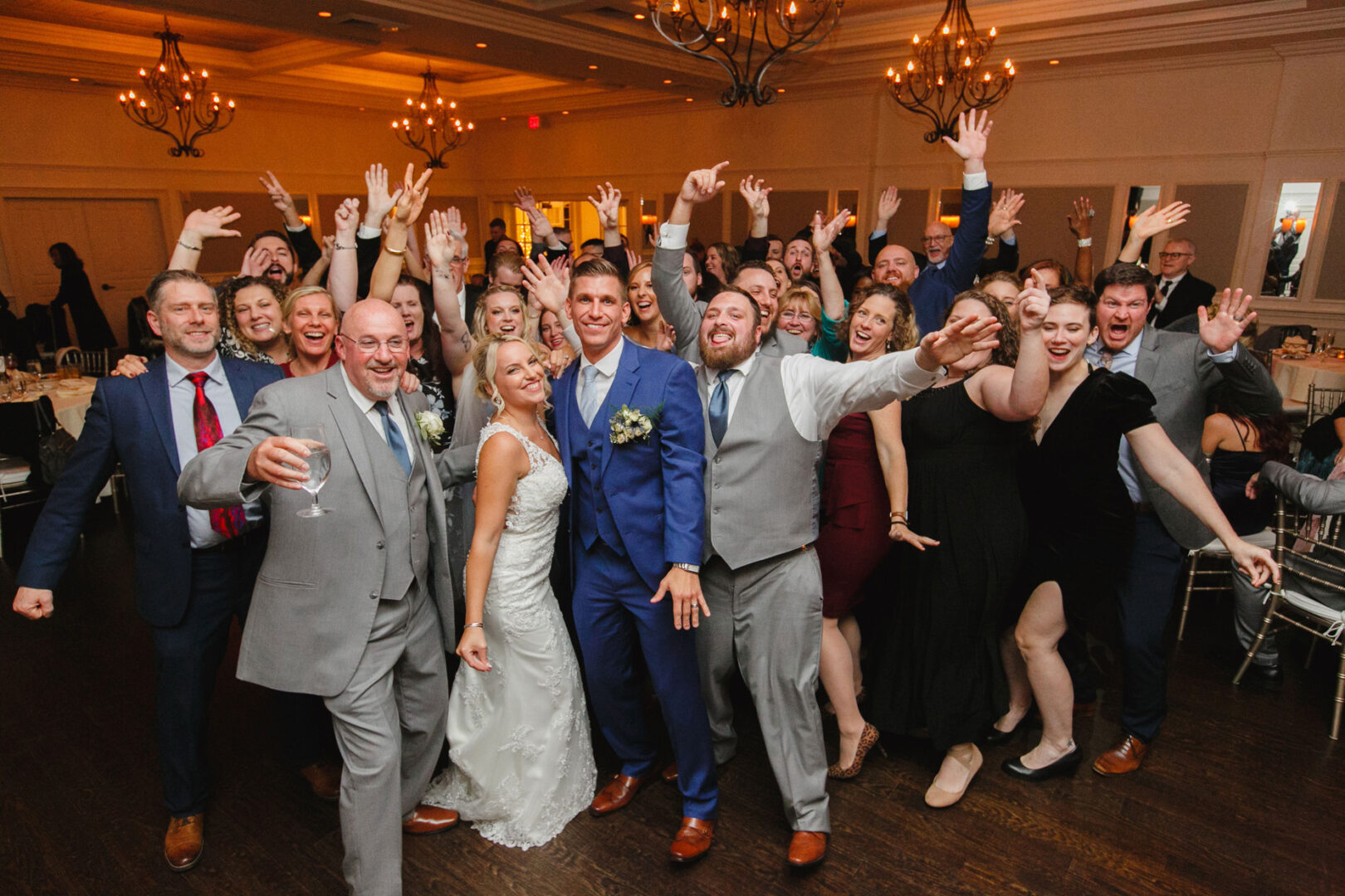 At the French Creek Golf Club wedding, a large group of people, including the joyful bride and groom, cheerfully pose with raised arms in the festive venue.