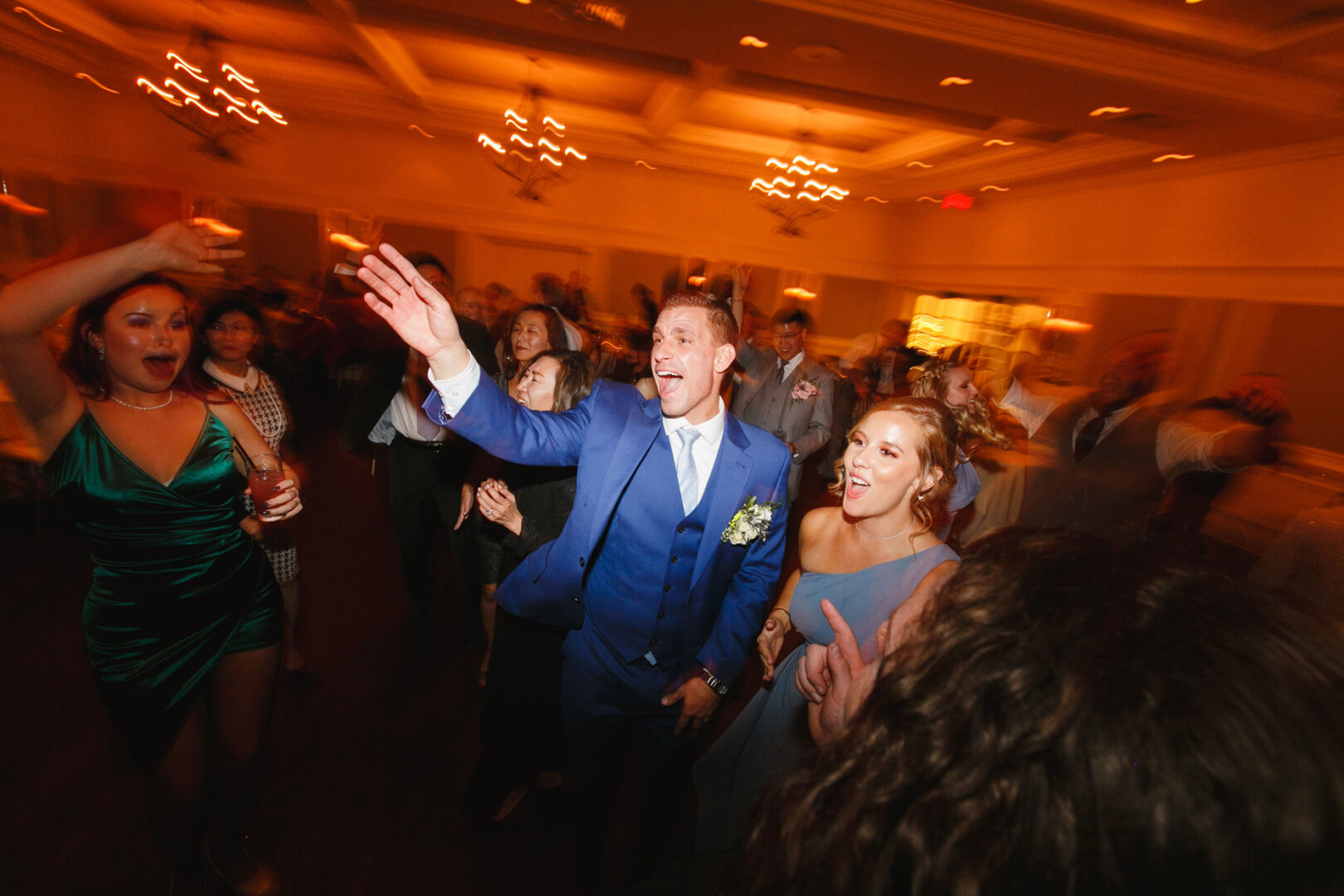People dancing energetically at a celebration in a warmly lit room at the French Creek Golf Club wedding. A man in a blue suit and a woman in a blue dress are prominent, smiling and reaching out.
