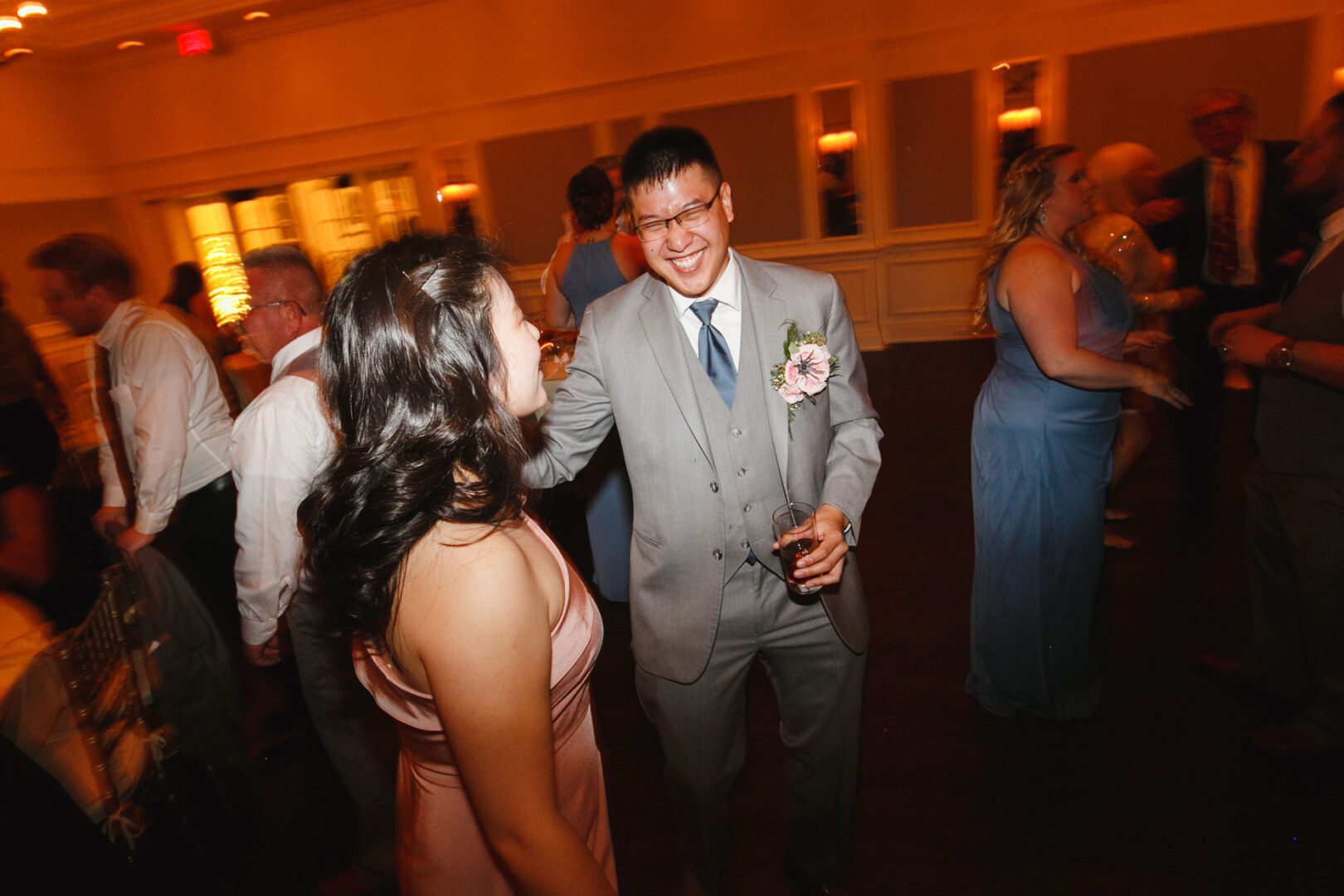 A man in a gray suit, holding a drink, smiles while chatting with a woman in a pink dress at the French Creek Golf Club Wedding. The social event buzzes with other elegantly dressed guests mingling in the background.