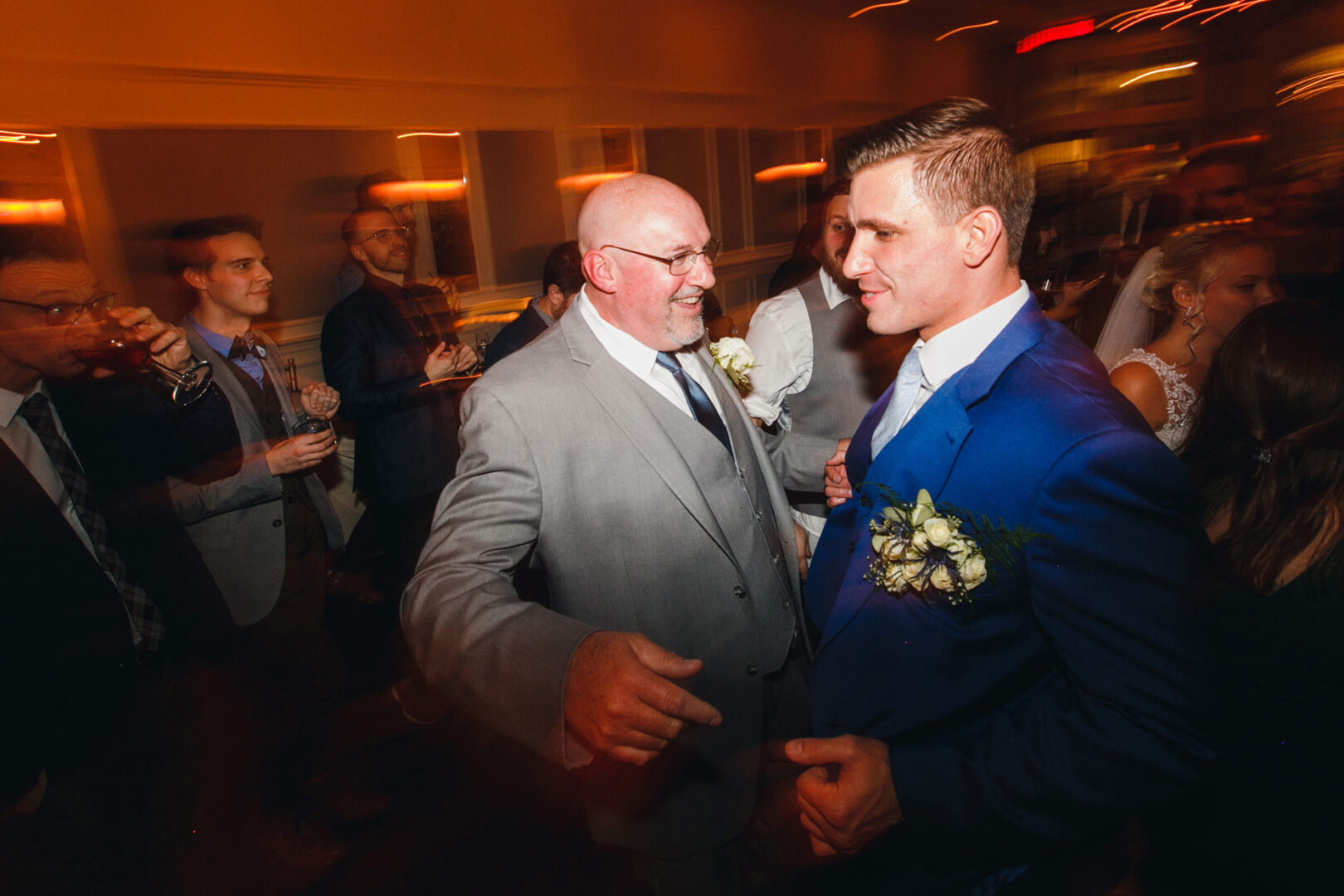 A man in a gray suit engages with a man in a blue suit at the French Creek Golf Club wedding reception. Guests mingle in the background, enhancing the scene's warm, lively atmosphere.