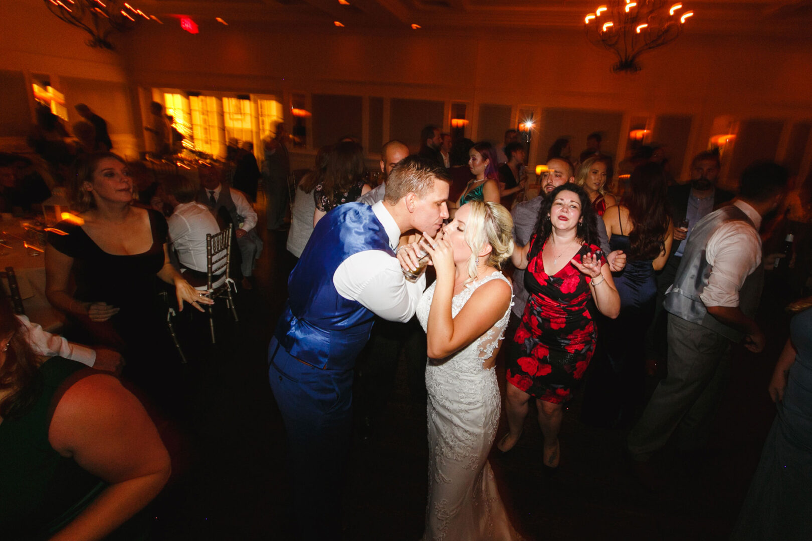 A bride and groom kiss on the dance floor at their enchanting French Creek Golf Club wedding reception, surrounded by guests in formal attire, with warm lighting and chandeliers above.