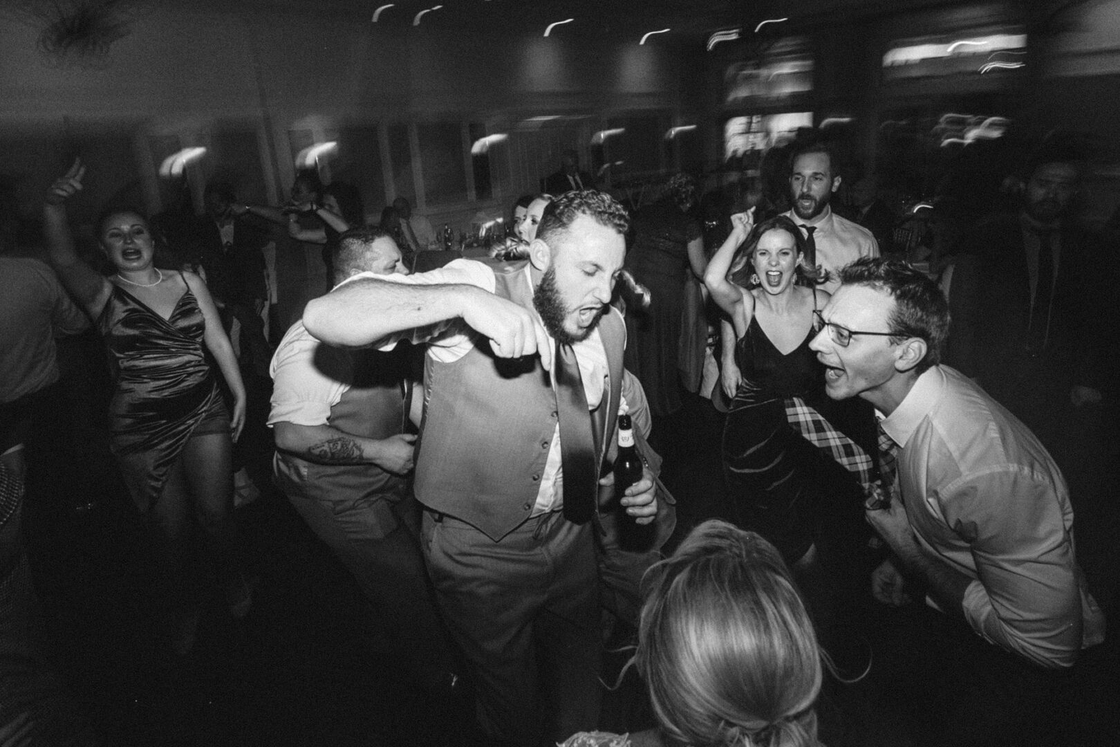 At a lively indoor event at French Creek Golf Club, a group of people energetically dance and sing, drinks in hand. The joyful, festive atmosphere is palpable with blurred motion capturing the wedding excitement.