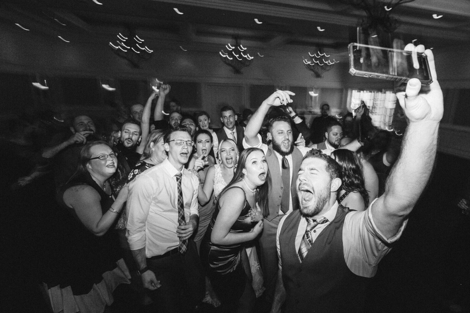 A group of people enthusiastically singing and cheering, with one person in the foreground taking a selfie. It's a lively indoor gathering at a French Creek Golf Club wedding.