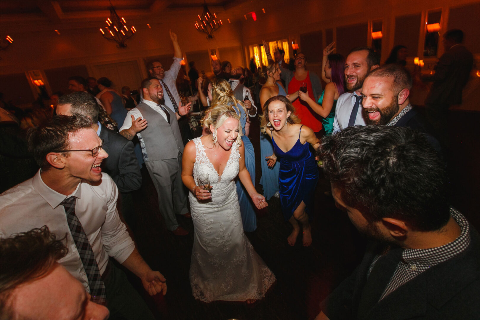 At a French Creek Golf Club wedding, a bride in a white dress celebrates with guests on the dance floor. Everyone is joyful and engaged, with some clapping and cheering amidst the vibrant atmosphere.