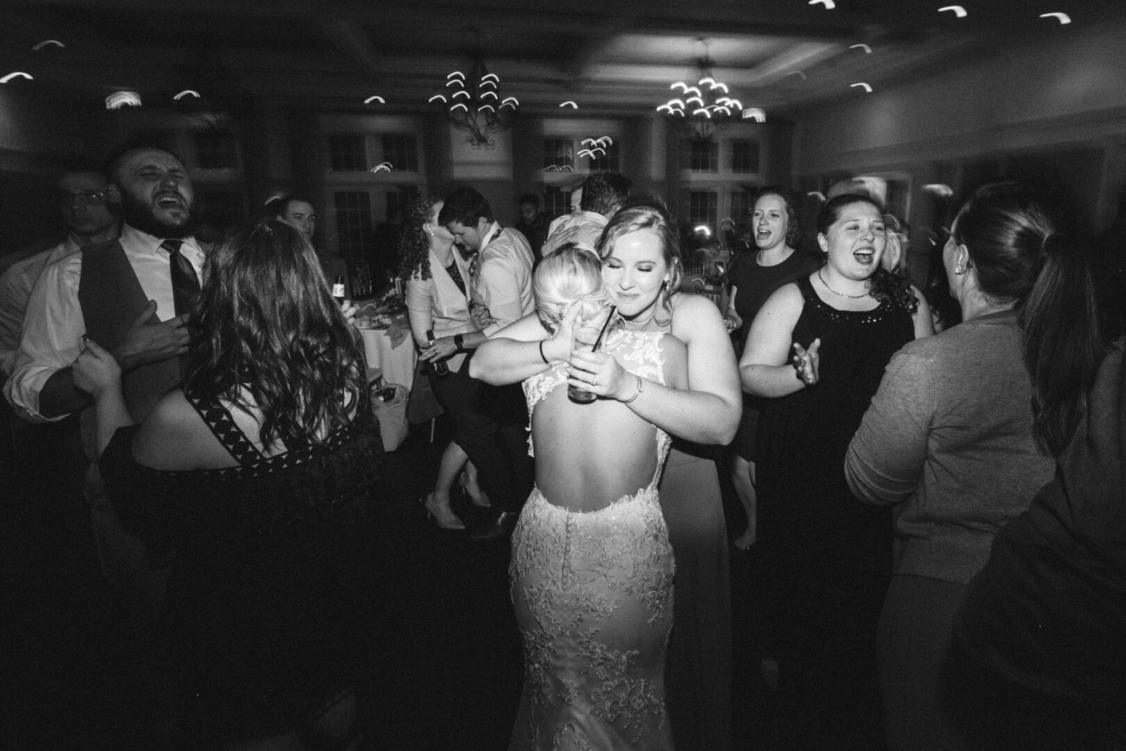 At a vibrant French Creek Golf Club wedding, the bride in a lace dress dances and hugs a guest on the lively ballroom floor, surrounded by people reveling in the joyous celebration.