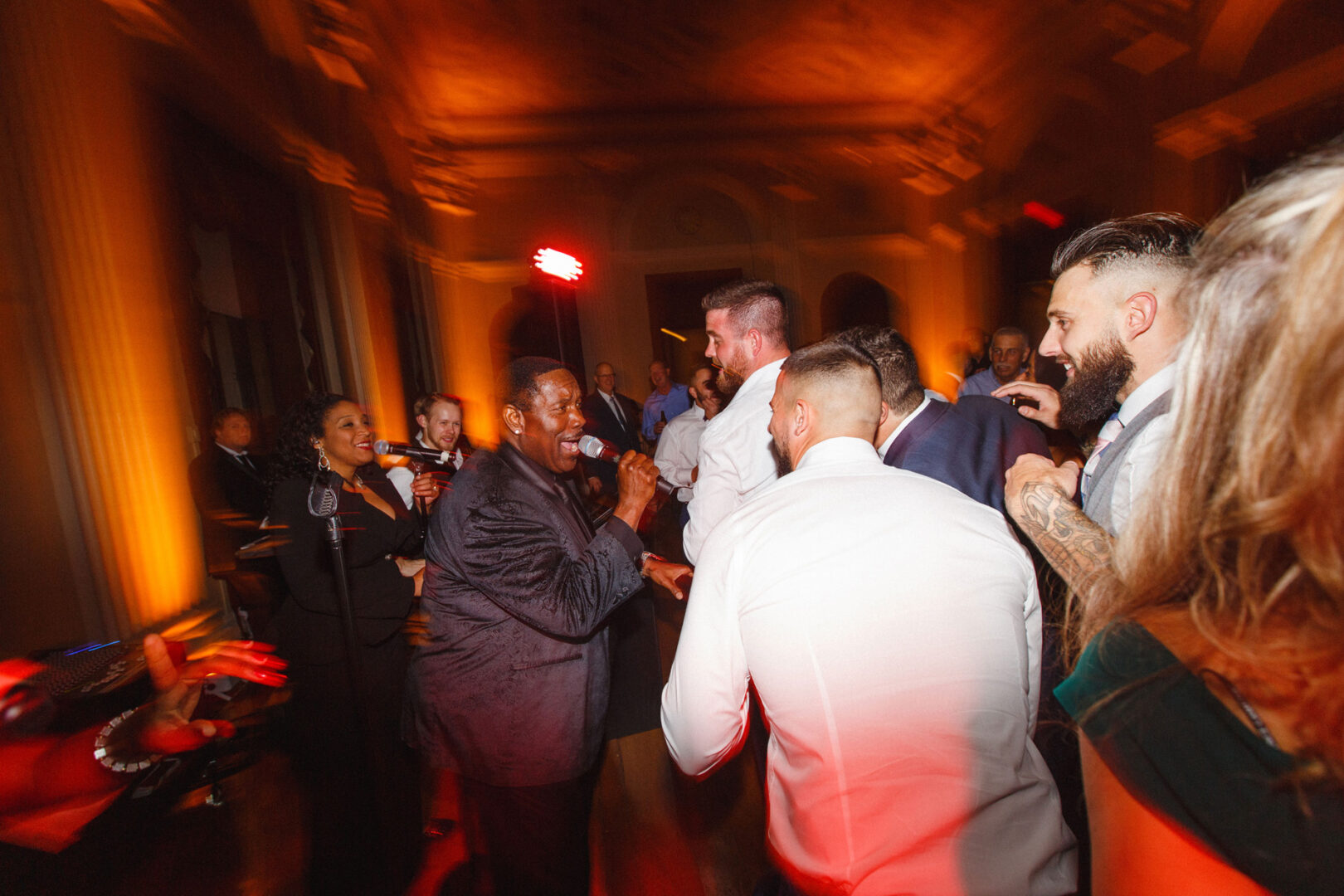 A lively indoor event unfolds at the Franklin Institute, with music and dancing filling the warmly lit room. One person passionately sings into a microphone, adding to the celebration.