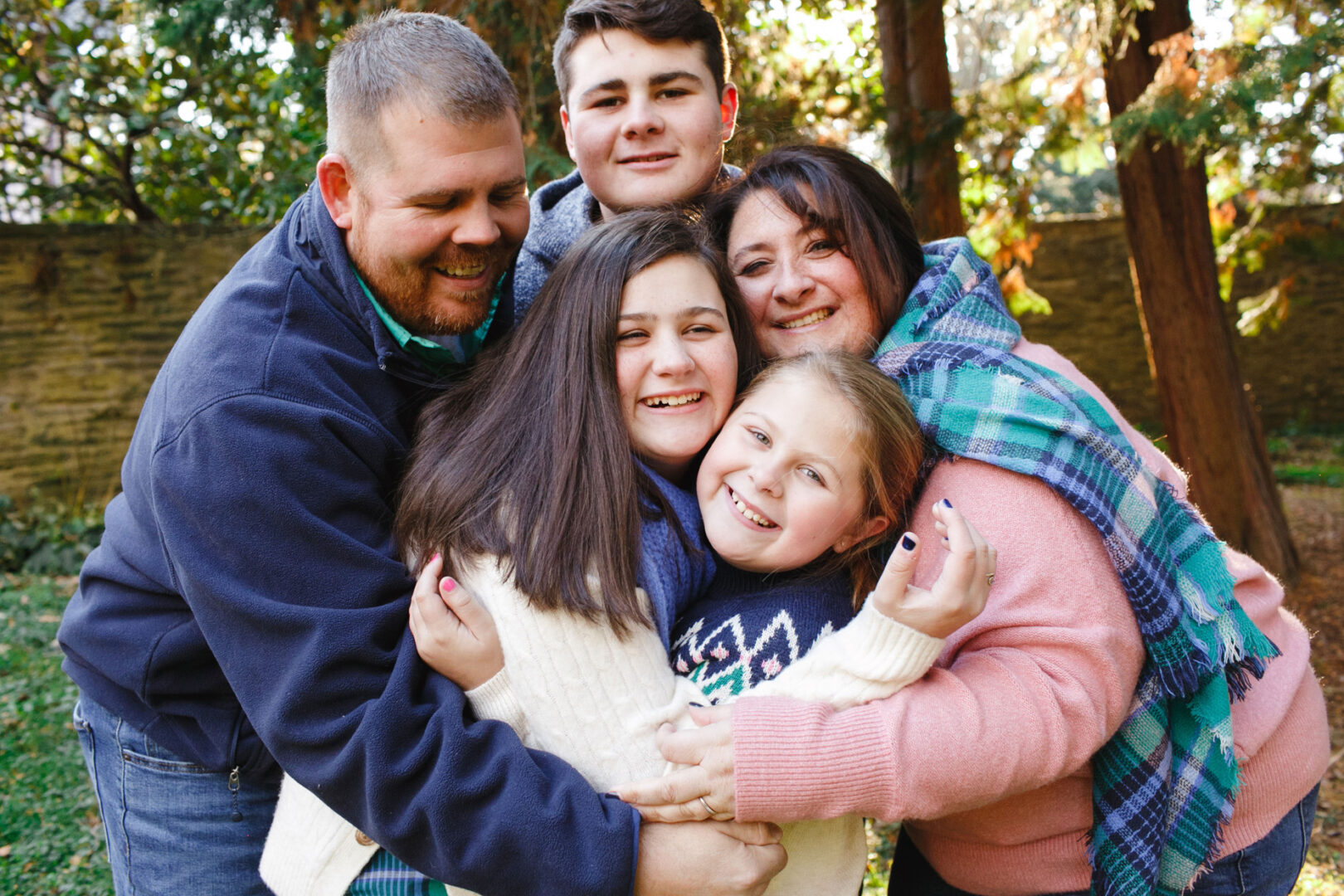 A family of five, including two adults and three children, pose closely together while smiling outdoors at Grange Estate. Trees and a stone wall add charm to the background of this heartwarming family photo.